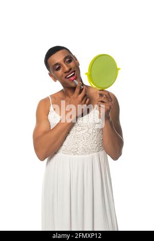 uomo travestito afroamericano in sundress che applica rossetto mentre osserva nello specchio isolato su bianco Foto Stock