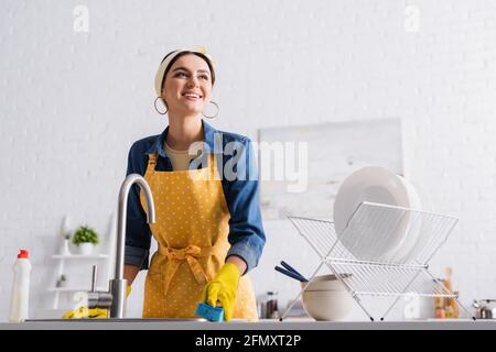 Sorridente casalinga in guanti di gomma che tengono la spugna vicino utensil e. lavello Foto Stock