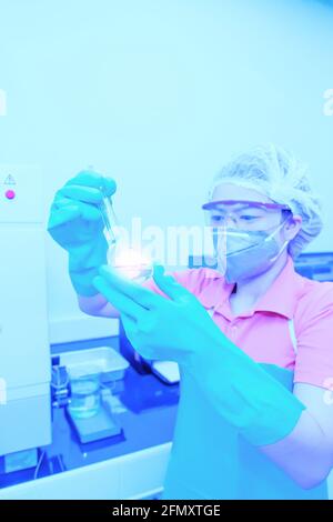 Una donna asiatica che indossa una maschera chirurgica che tiene un matraccio volumetrico o una provetta che fa la ricerca in un laboratorio. Foto Stock