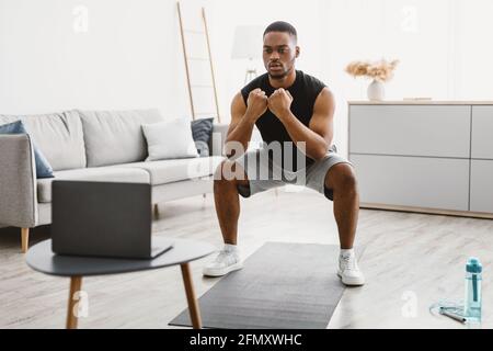Atletico afroamericano Guy facendo squats al laptop a casa Foto Stock