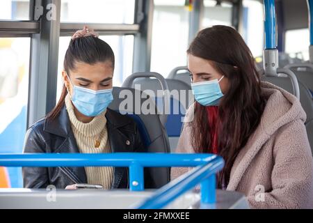 Due giovani donne negli anni 20 seduti all'interno di un autobus indossare maschere e guardare un telefono cellulare in le loro mani Foto Stock
