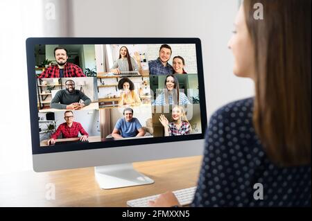 Conferenza, riunione mattutina online. Una giovane donna sta usando il laptop per la connessione con diversi colleghi, dipendenti multirazziali. Videochiama con molte persone insieme. Vista posteriore Foto Stock