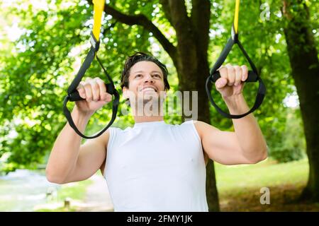 Giovane uomo che esercitano con sospensione trainer fionda in City Park in estate gli alberi per lo sport fitness Foto Stock