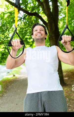 Giovane uomo che esercitano con sospensione trainer fionda in City Park in estate gli alberi per lo sport fitness Foto Stock