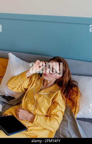Una giovane donna con un comodo pigiama giallo lavora mentre si sdraia il letto in camera da letto. una donna d'affari mantiene un distanziamento sociale lavora a distanza. Nuovo concetto normale Foto Stock