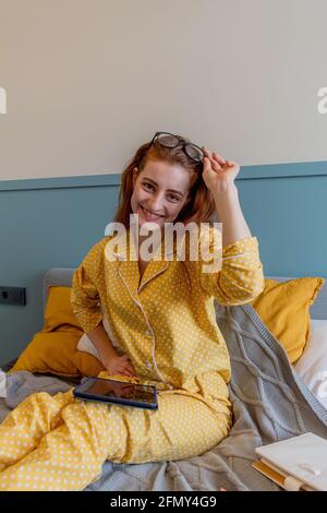 Una giovane donna con un comodo pigiama giallo lavora mentre si sdraia il letto in camera da letto. una donna d'affari mantiene un distanziamento sociale lavora a distanza. Nuovo concetto normale Foto Stock