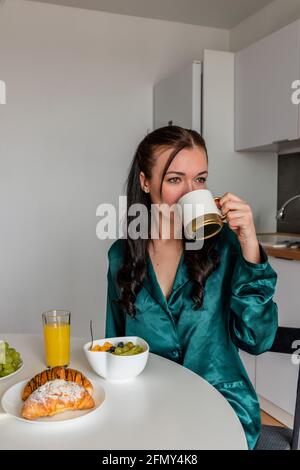 Giovane donna di seta pigiama ha la prima colazione e bevande caffè in cucina a casa. Concetto di benessere. Foto Stock