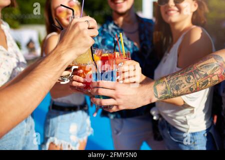 Vista ravvicinata degli amici che si divertono al party estivo a bordo piscina, bicchieri da clinking con cocktail estivi freschi vicino alla piscina all'aperto dell'hotel. Persone Foto Stock