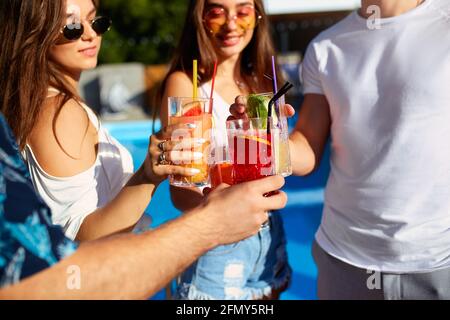 Vista ravvicinata degli amici che si divertono al party estivo a bordo piscina, bicchieri da clinking con cocktail estivi freschi vicino alla piscina all'aperto dell'hotel. Persone Foto Stock