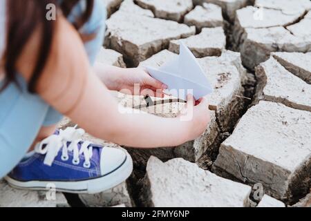 La ragazza abbassa la barca di carta sul terreno asciutto e incrinato. Crisi idrica e concetto di cambiamento climatico. Riscaldamento globale Foto Stock