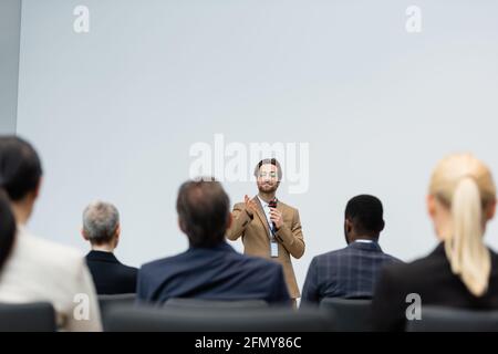 Altoparlante sorridente con microfono che punta con la mano vicino a un'azienda multietnica persone Foto Stock