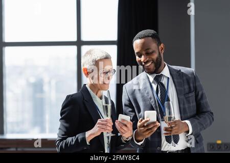 partner commerciali interrazziali felici con champagne e telefoni cellulari che parlano durante conferenza aziendale Foto Stock