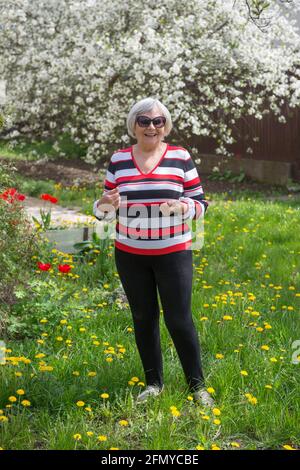 Ritratto a lunghezza intera di donna anziana sorridente che sta in piedi su prato verde luminoso al suo cortile con ciliegio fiorente. Foto Stock