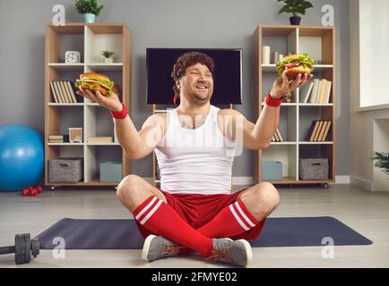 Uomo millenario eccitato con corpo chubby che tiene hamburger fast food in due mani Foto Stock