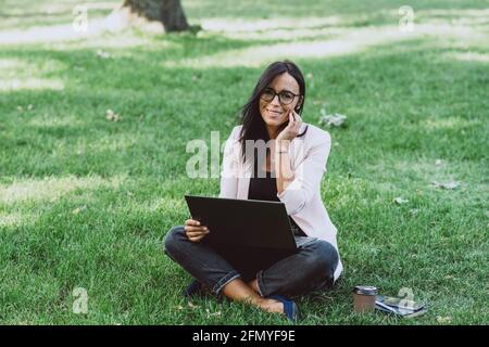 Donna d'affari che siede nel parco estivo dell'erba usando il laptop. Un uomo d'affari lavora a distanza durante la quarantena. All'aperto. Messa a fuoco selettiva morbida. Foto Stock