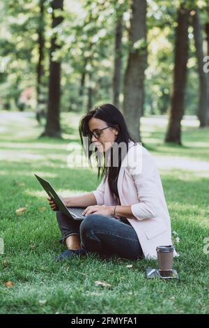 Donna d'affari che siede nel parco estivo dell'erba usando il laptop. Un uomo d'affari lavora a distanza durante la quarantena. All'aperto. Messa a fuoco selettiva morbida. Foto Stock