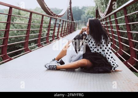 Una bella giovane donna in una gonna soffice e una blusa polka-dot si siede sul ponte e bacia il suo cane Staffordshire Bull Terrier. Focu soft selective Foto Stock