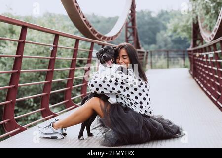 Una bella giovane donna in una gonna soffice e una blusa polka-dot si siede sul ponte e abbraccia il suo cane Staffordshire Bull Terrier. Messa a fuoco selettiva morbida. Foto Stock