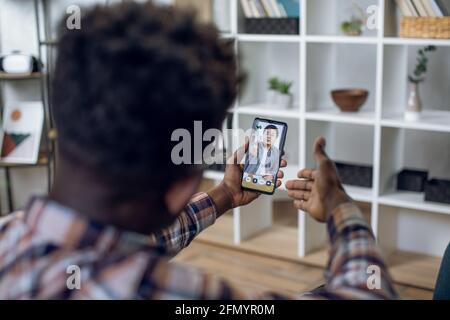 Giovane uomo nero seduto a casa e utilizzando il moderno smartphone per l'intervista di lavoro online con il datore di lavoro asiatico. Concetto di lavoro remoto, persone e tecnologia. Foto Stock