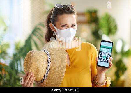 Viaggio durante la pandemia del coronavirus. Ritratto di felice elegante donna turistica con smartphone, covid app passaporto, maschera medica e cappello di paglia nel mod Foto Stock