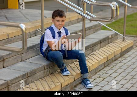 Scolaro in una camicia bianca con una giacca blu si siede sulle scale e gioca con un tablet grigio Foto Stock