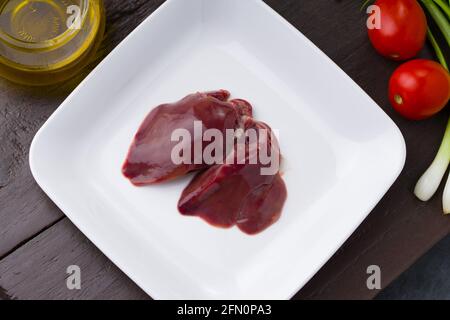 Fegato di pollo crudo disposto splendidamente in un piatto bianco di ceramica con pomodoro piccolo, cipolla primaverile e olio da cucina su sfondo Foto Stock