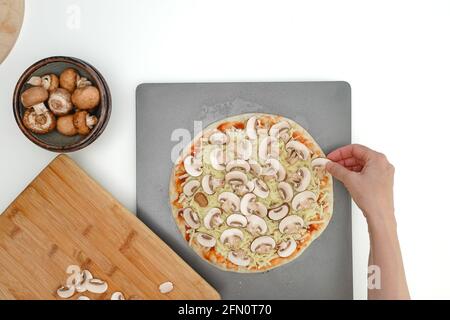 Affettare le fette di funghi dell'impasto della pizza, vista ravvicinata, posa piatta, mani femminili. Pizza fatta in casa con mozzarella e funghi, ricetta passo dopo passo Foto Stock
