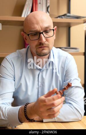 uomo d'affari calvo con gli occhiali guarda pensieroso al pezzo di scacchi in mano. Sviluppare una strategia, prendere decisioni importanti Foto Stock