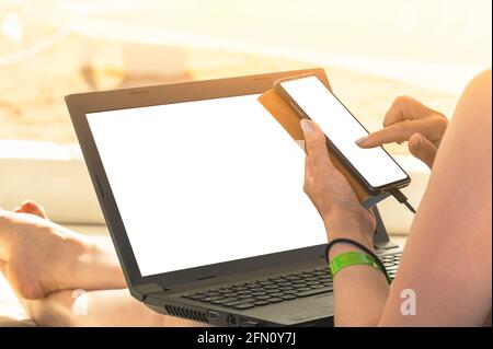 Mockup di computer portatile e telefono cellulare nelle mani di una donna. Donna d'affari freelance che lavora sulla spiaggia utilizzando computer portatile e telefono cellulare. Stile di vita per le vacanze Foto Stock