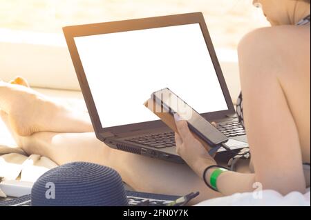 Immagine mockup. Donna che usa un computer portatile con schermo bianco vuoto e tiene un telefono cellulare su una spiaggia durante le vacanze. Lavoratrice freelance Foto Stock