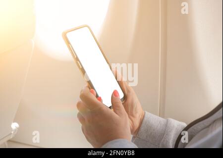 Donna che usa lo smartphone in aereo. Immagine closeup di donna che tiene e tocca mockup cellulare accanto a una finestra di aereo. Mano femmina che tiene SMA Foto Stock
