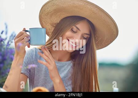 Fuoco selettivo della ragazza giovane che indossa il cappello grande di paglia e vestito che tiene la bevanda calda all'aperto. Splendida donna caucasica che beve caffè, in posa in campo di lavanda. Concetto di bellezza, natura, bevanda. Foto Stock