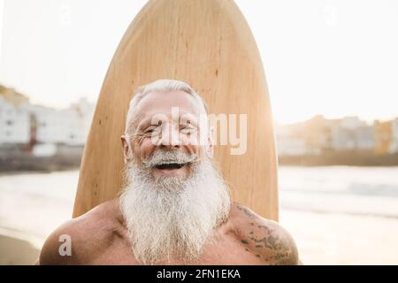 Surfista senior tatuato che tiene una tavola da surf vintage sulla spiaggia Al tramonto - fuoco sul viso Foto Stock