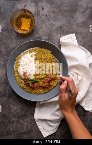 Risotto cremoso di fave con burrata (piatto tipico pugliese) e prosciutto italiano croccante. Fondo grigio, piastra grigia e asciugamano bianco Foto Stock