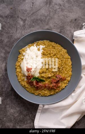 Risotto cremoso di fave con burrata (piatto tipico pugliese) e prosciutto italiano croccante. Fondo grigio, piastra grigia e asciugamano bianco Foto Stock