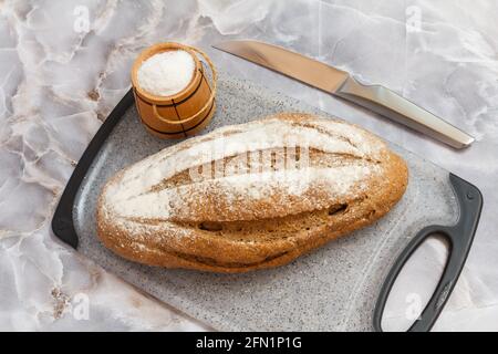 Pane di segale e una cantina di sale di legno su un tagliere, un coltello da cucina con sfondo grigio. Vista dall'alto. Foto Stock