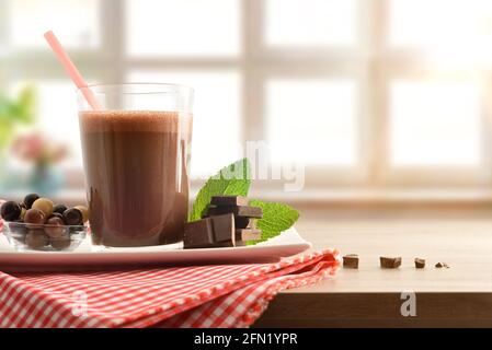 Bicchiere con shake di cioccolato al latte sul piatto di portata con recipiente pieno di palline di colato e porzioni di colore rosso e bianco tovaglia a scacchi su un tavolo con l Foto Stock