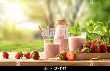 Frullato di fragole con latte in contenitori di vetro su tavola di legno con vassoio pieno di fragole in natura. Vista frontale. Foto Stock