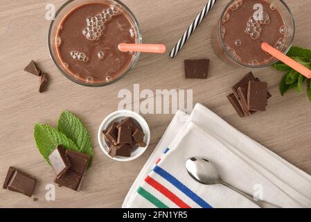 Due bicchieri con frullato di cioccolato al latte e porzioni di cioccolato su panca di legno. Vista dall'alto. Foto Stock