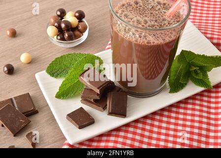 Bicchiere con frullato di cioccolato al latte sul piatto con palle e porzioni di cioccolato su tovaglia a scacchi rosso e bianco su panca di legno. Vista rialzata. Foto Stock