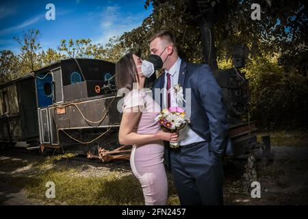 Coppie di nozze in maschere mediche pone nel parco. Newlyweds durante la pandemia di Coronavirus nelle maschere facciali. Sposo e sposa si baciano in protectiv Foto Stock