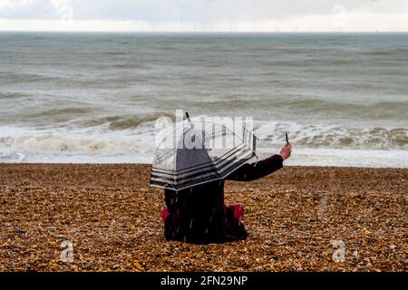 Una giovane donna seduta nella pioggia sulla spiaggia di Brighton prendendo UN Selfie, Brighton, Sussex orientale, Regno Unito. Foto Stock