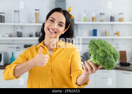 donna bruna felice che tiene broccoli verde maturo mentre mostra il pollice su Foto Stock