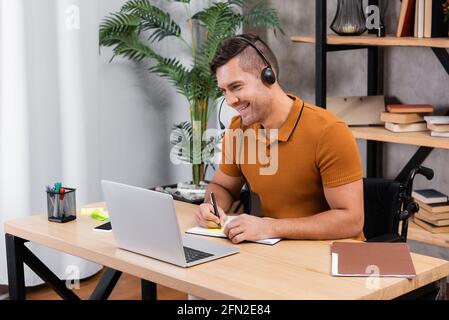 uomo sorridente e disabile che scrive nel notebook mentre lavora in cuffia a casa Foto Stock