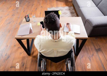 vista dall'alto di un uomo portatore di handicap che tiene il cellulare e che scrive notebook vicino al computer portatile Foto Stock