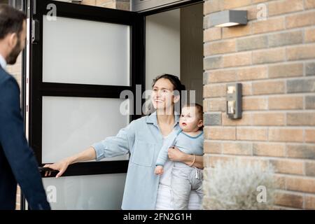 Casalinga con neonato che si trova di fronte alla porta d'ingresso della casa e saluta un marito e un papà dal lavoro. Foto Stock