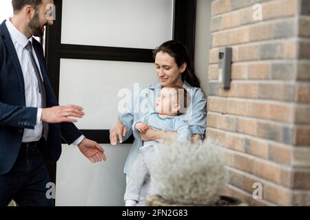 Casalinga con neonato che si trova di fronte alla porta d'ingresso della casa e saluta un marito e un papà dal lavoro. Foto Stock