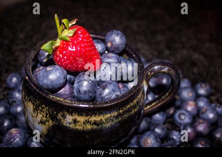 Una sola fragola rossa luminosa sopra una tazza da tè nera, ripiena di grandi bacche blu di colore indaco Foto Stock