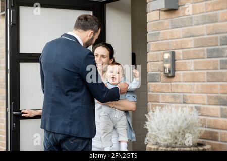 Casalinga con neonato che si trova di fronte alla porta d'ingresso della casa e saluta un marito e un papà dal lavoro. Foto Stock