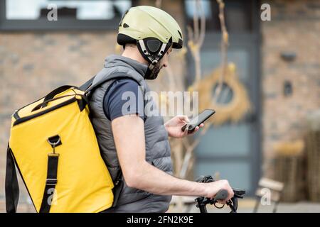 L'addetto alla consegna in casco con zaino giallo guida uno scooter elettrico per strada e cerca per telefono l'indirizzo del cliente. Foto Stock